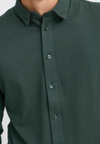 Camisa de botones de color verde oscuro hecha de tela texturizada, con cuello y cinco botones a juego en la parte delantera.