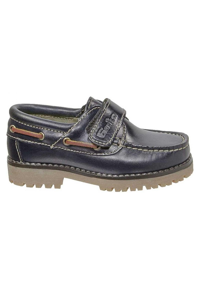 Zapato slip-on de cuero azul marino con una correa de velcro, suela de goma con ranuras de tracción, y costuras y acentos en contraste en color marrón claro.