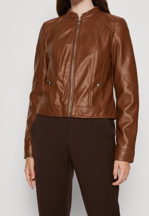 Kvinde iført en tætsiddende brun læderjakke med lynlås foran og lommedetaljer med trykknapper, kombineret med mørkebrune bukser.