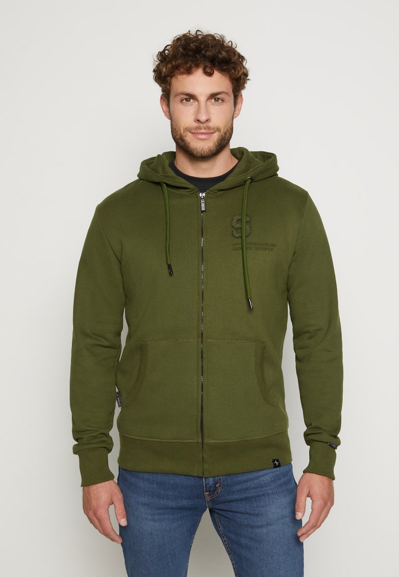 Olivgrüner Zip-Hoodie mit Kapuze und Zugband, Fronttaschen und einem strukturierten Logo auf der Brust. Der Stoff ist weich und hat gerippte Bündchen.