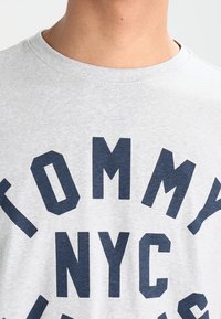 Harmaa t-paita, joka on valmistettu puuvillasta, ja jonka etupuolella on suuri tummansininen teksti "TOMMY NYC" rohkealla, isoilla kirjaimilla. Pyöreä kaula-aukko.