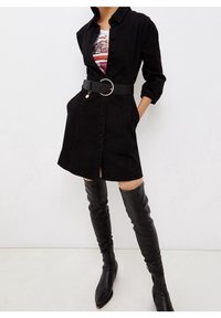 Abito camicia in denim nero con bottoni frontali, colletto e maniche arrotolate, cinte con una cintura nera con un dettaglio di perle. Abbinato a stivali neri alti fino alla coscia.