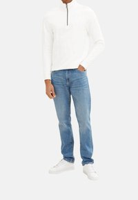 Sudadera blanca con cremallera y mangas largas, combinada con jeans de color azul claro y zapatillas blancas. La sudadera tiene una superficie texturizada.