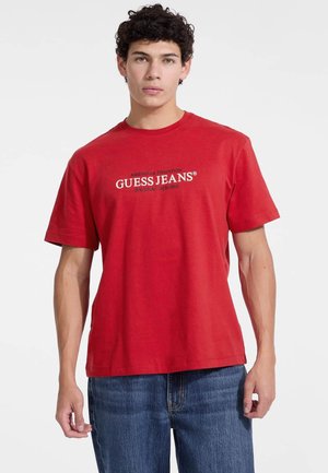Tricou roșu din bumbac cu mâneci scurte, având logo-ul "GUESS JEANS" inscripționat cu text alb pe piept. Guler rotund și croială lejeră.