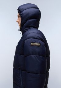 Mornarska puhovka s kapuco, z odebeljenim dizajnom, z zadrgo na stranski žepi in slogovno oznako na levem rokavu. Gladka tekstura.