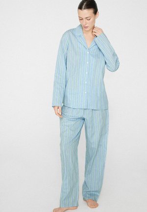 Femme portant un haut de pyjama à rayures bleu clair à manches longues avec boutons et un pantalon ample assorti, debout pieds nus devant un fond uni.