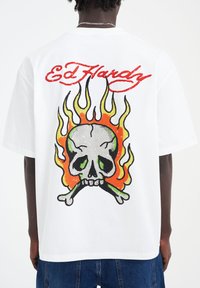 White t-shirt featuring a large color graphic of a skull with green details, crossbones, and orange flames, with "Ed Hardy" in red above.
