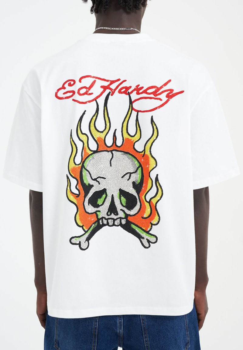 White t-shirt featuring a large color graphic of a skull with green details, crossbones, and orange flames, with "Ed Hardy" in red above.