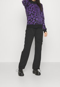 Lila und schwarz gemusterter Pullover mit Rundhalsausschnitt, kombiniert mit schwarzen Cargo-Hosen mit Seitentaschen und schwarzen Schnürschuhen.