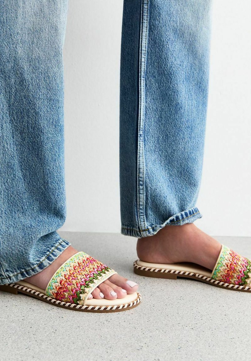 Las sandalias deslizantes tejidas multicolores cuentan con un borde trenzado, suela plana y plantilla suave, combinadas con unos jeans de pierna recta en azul claro.