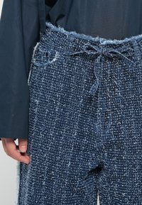 Dark blue, textured fabric trousers with a frayed waistband and drawstring. Features a loose fit and distressed details.
