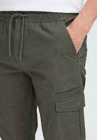 Olivgrüne Cargo-Hose mit elastischem Bund, Kordelzugverschluss, Seitentaschen und einer Klapptasche am Oberschenkel. Aus Baumwollmaterial.