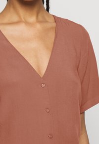 Haut couleur rouille avec un col en V, des manches courtes et cinq boutons sur le devant. Confectionné dans un tissu doux et texturé.