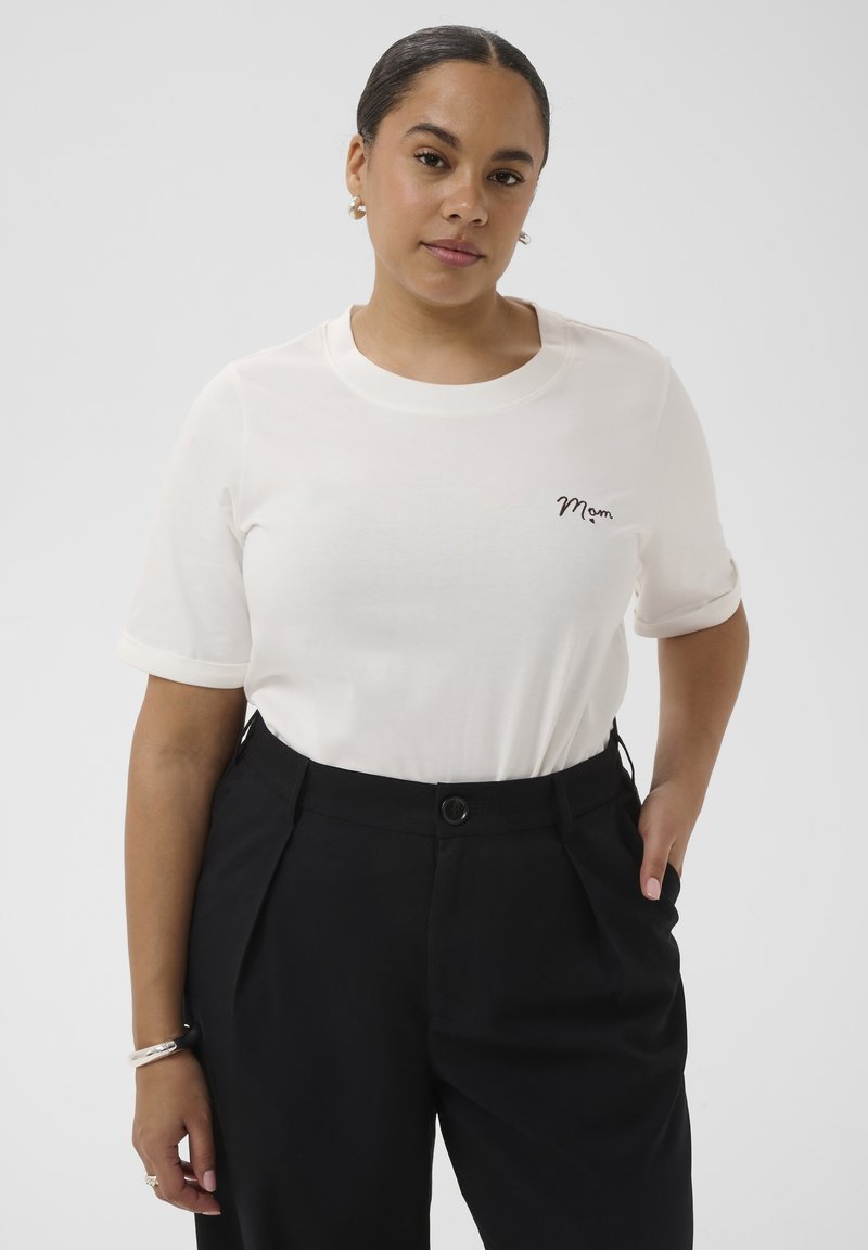 Femme aux cheveux noirs portant un t-shirt blanc avec l'inscription "Mom" sur la poitrine, un pantalon noir taille haute, debout avec une main dans la poche devant un fond uni.