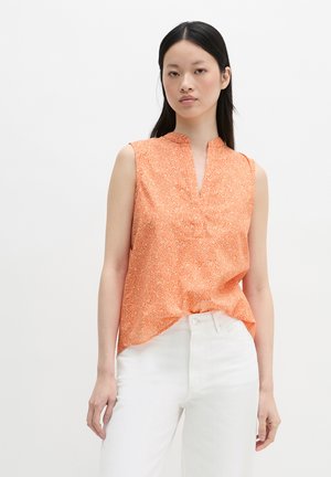 Femme portant un haut orange sans manches à motifs floraux avec un col en V et un pantalon blanc, debout devant un fond blanc uni.