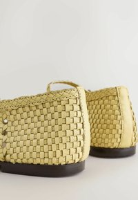 Chaussures plates en cuir tressé de couleur jaune clair avec bordure tressée et semelles foncées, vues de dos sur un fond blanc.