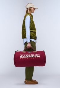 Borsa a sacca color bordeaux con manici in pelle marrone, con logo e dettaglio di piccola bandiera. Il modello indossa un gilet verde imbottito e un berretto beige.