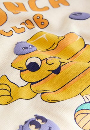 Piirrosjuustohahmo suurilla silmillä ja kielen ulkona, jota ympäröivät hymyilevät violetit marjat kuvioidulla valkoisella kankaalla.