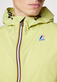 Jasnozielona wodoodporna kurtka z kapturem, z przednim zamkiem błyskawicznym zdobionym niebieskimi, pomarańczowymi i białymi paskami oraz logo K-Way na piersi.