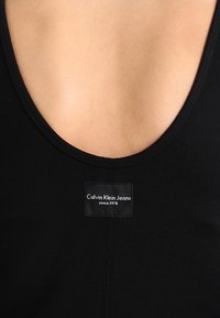 Nahaufnahme eines schwarzen Calvin Klein Jeans Kleidungsstücks mit U-Boot-Ausschnitt, das nackte Haut zeigt, und einem aufgenähten Markenlabel.