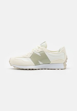 Seitenansicht eines hellbeigen und weißen Sneakers mit einem großen grauen Buchstaben "N" an der Seite und weißen Schnürsenkeln auf einfarbigem Hintergrund.