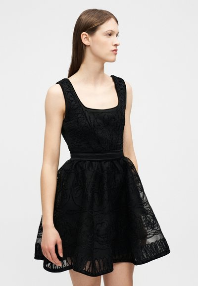 Νεαρή γυναίκα που φοράει μαύρο δαντελένιο φόρεμα χωρίς μανίκια με εφαρμοστή μπλούζα και φουσκωτή φούστα, στέκεται μπροστά σε μια λευκή απλή επιφάνεια.