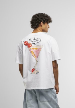 Young man with curly hair wearing a white T-shirt featuring a pink cocktail glass with cherries and French text on the back.