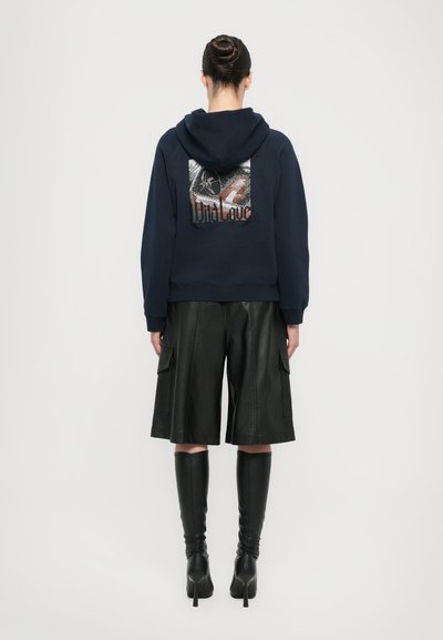 Marineblaues Kapuzensweatshirt mit Grafikdruck auf der Rückseite, kombiniert mit schwarzen Ledershorts und kniehohen Stiefeln. Glatte Textur, lässiges Design.