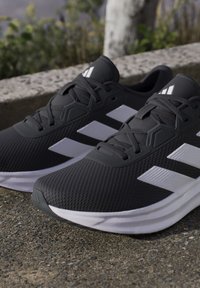 Paire de chaussures de course noires avec des bandes blanches et des lacets placées sur une surface en béton en extérieur près des plantes.