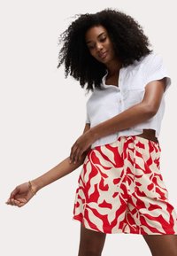Weißes, kurzes Hemd mit Knöpfen. Rote und cremefarbene gemusterte Shorts mit Kordelzug. Fokus auf Blumenmuster und entspannte Passform.