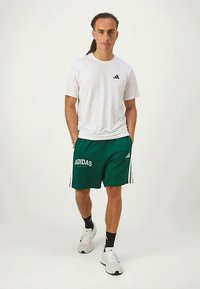 Biela krátkopiesková košeľa s čiernym logom Adidas; zelené šortky s bielym textom "ADIDAS"; biele športové topánky s čiernymi ponožkami.