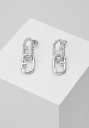Boucles d'oreilles créoles en argent avec deux formes ovales reliées, l'une incrustée de petits cristaux clairs et l'autre en métal lisse, présentées sur un bloc blanc.
