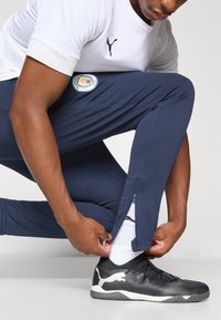 Pantalons de sport bleu marine en tissu texturé, avec des ouvertures zippées sur les côtés et un logo de Manchester City. Portés avec un t-shirt en mesh blanc et des baskets noires.