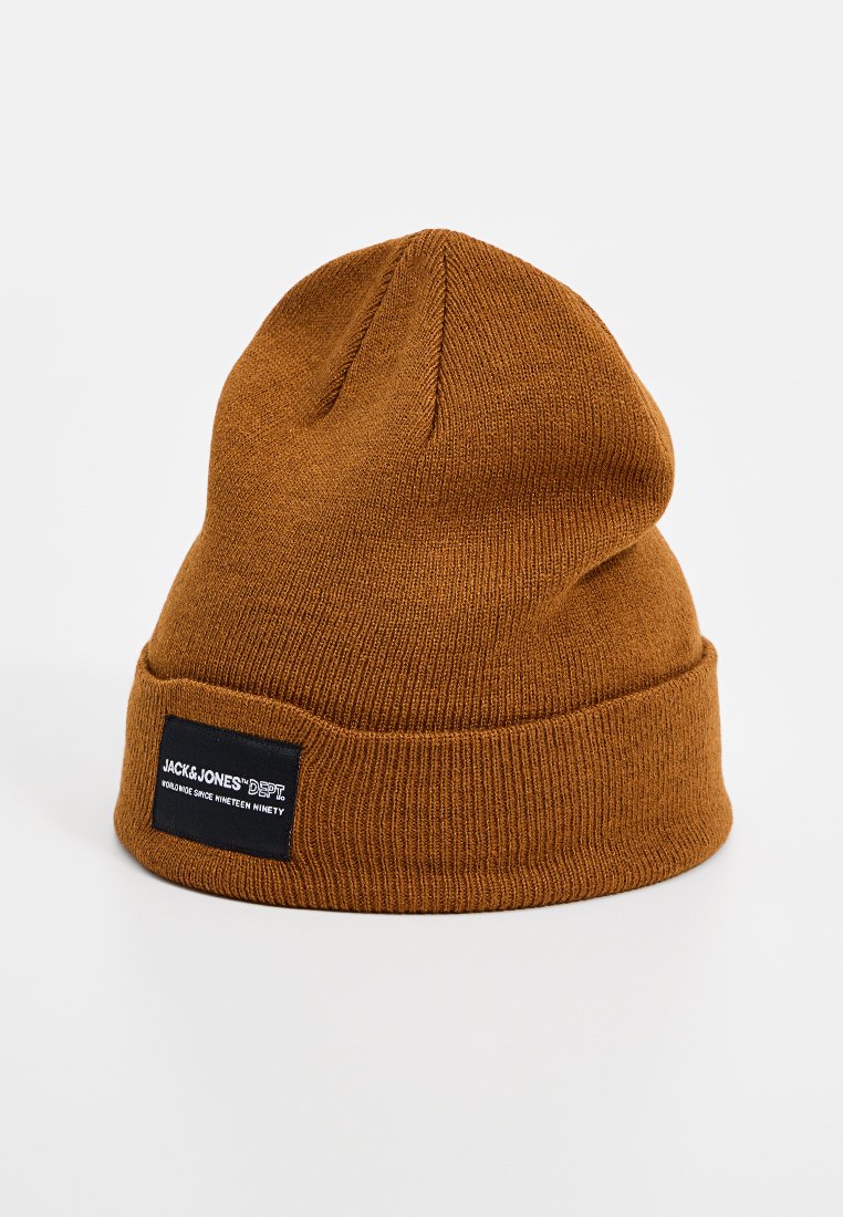 Bruine gebreide beanie muts met een omgeslagen rand, voorzien van een rechthoekig zwart label met witte tekst. Zachte textuur, eenvoudig ontwerp.