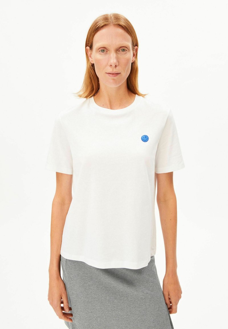 Weißes Baumwoll-T-Shirt mit Rundhalsausschnitt, versehen mit einem kleinen blauen Smiley-Emblem auf der linken Brust. Kurze Ärmel und entspannte Passform.