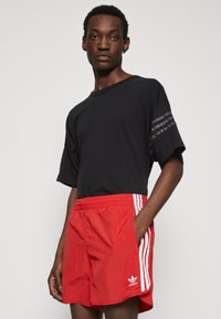 Tricou negru cu mânecă scurtă, având o dungi texturate pe umăr, asociat cu pantaloni scurți roșii vibranți, cu dungi albe pe laterale și un logo.