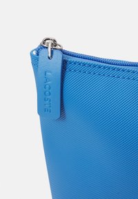 Pochette zippée texturée bleue avec une tirette en cuir embossée du nom de la marque "LACOSTE".