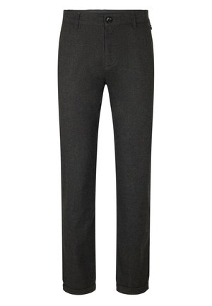 Mann trägt einen schwarzen Pullover mit Viertel-Reißverschluss, dunkelgraue Hosen mit Bündchen, weiße Socken und weiße Sneakers, steht mit den Händen hinter dem Rücken.
