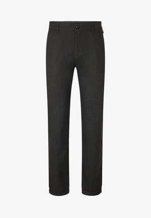 Mand iført en sort kvart-lynlåstrøje, mørkegrå bukser med manchetter, hvide sokker og hvide sneakers, stående med hænderne bag ryggen.