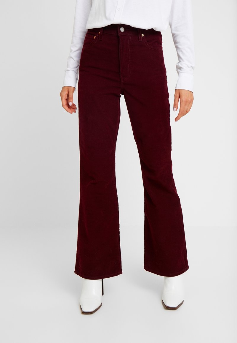 Pantalon évasé en velours côtelé bordeaux avec poches avant, taille haute et garnitures dorées, associé à des bottines blanches à bouts pointus.