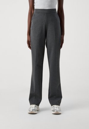 Osoba nosząca ciemnoszare spodnie z prostymi nogawkami, białe i srebrne tenisówki oraz jasnoszary bezrękawnik, stojąca na jednolitym tle.