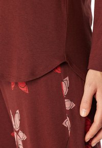 Camiseta de manga larga color marrón burdeos con un dobladillo curvado, combinada con pantalones estampados que presentan diseños florales rojos y una textura textil suave.