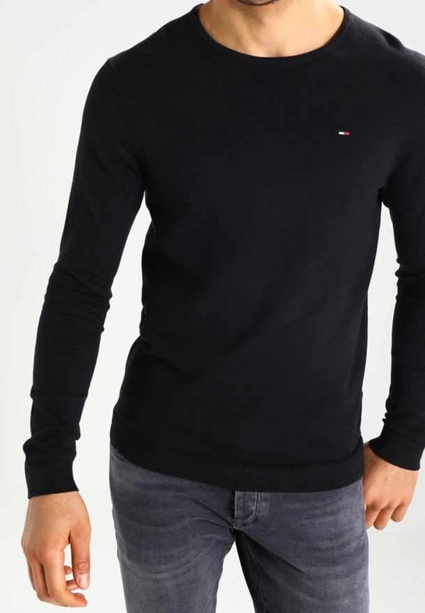 Pull en maille noir à manches longues avec un col rond, des poignets et un ourlet côtelés ; présente un petit logo sur la poitrine. Porté avec un jean gris.