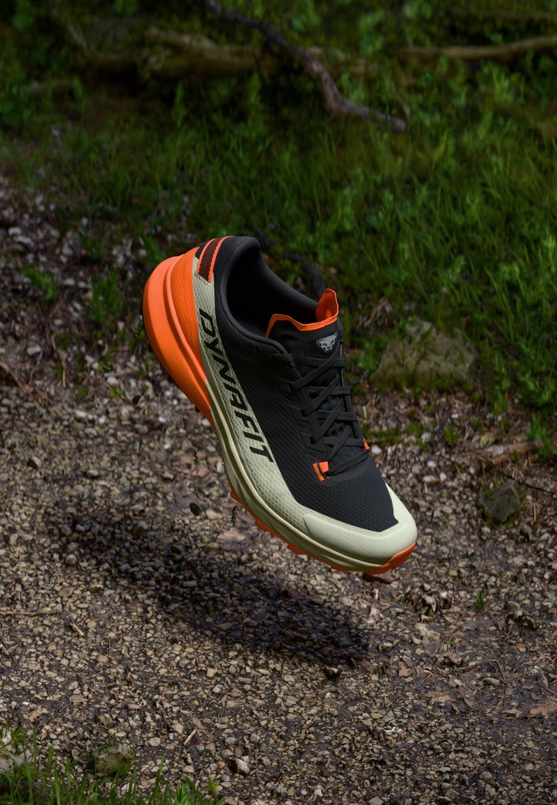 Chaussure de sport noire avec des accents orange et une semelle beige, présentant une tige texturée, des lacets et un logo sur le côté, flottant au-dessus d'un chemin de gravier.