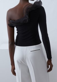 Top nero a spalla unica con dettaglio di rouches trasparenti; manica lunga; abbinato a pantaloni bianchi con una tasca accentata in nero.