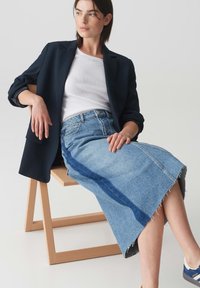 Denim-Midi-Rock mit fransigem Saum, mit seitlichem Panel aus dunklerem blauen Denim, getragen mit einem enganliegenden weißen Oberteil und einem marineblauen Blazer.