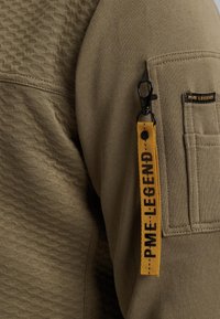 Khaki lange mouw top van textuurstof. Heeft een gele label met zwarte tekst en een clip op de mouwenzak voor extra detail.