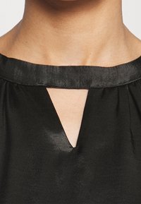 Černá blůza s saténovým povrchem, má kulatý výstřih s V-vykrojením, plisované detaily a hladkou strukturu podél okrajů.