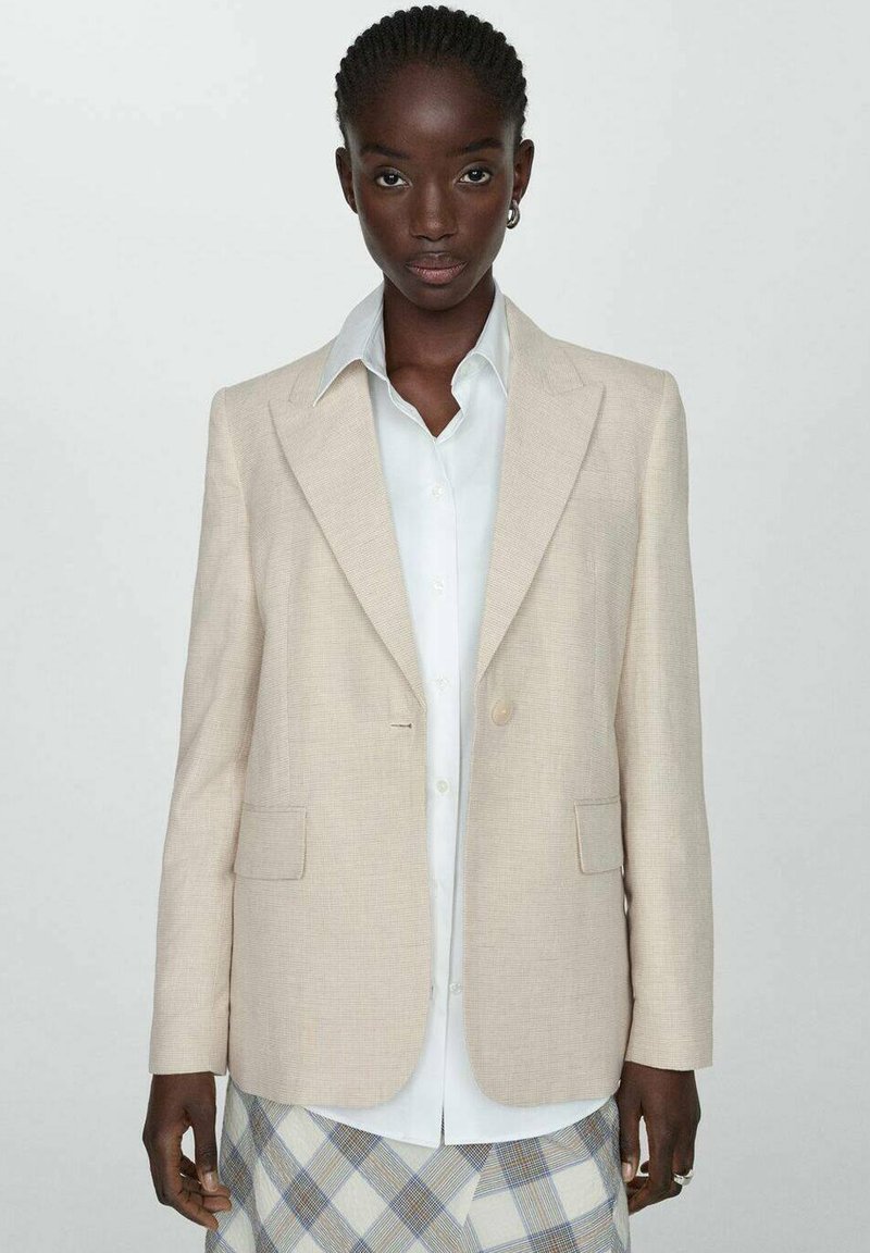 Beige Blazer aus strukturiertem Material, mit einem einzigen Knopfverschluss, Reverskragen und zwei vorderen Taschen, getragen über einem weißen Hemd.
