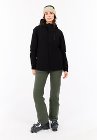 Vrouw staat op een witte achtergrond, gekleed in een beige gebreide beanie, een zwarte hooded jacket, olijfgroene broek en zwarte skibotten.